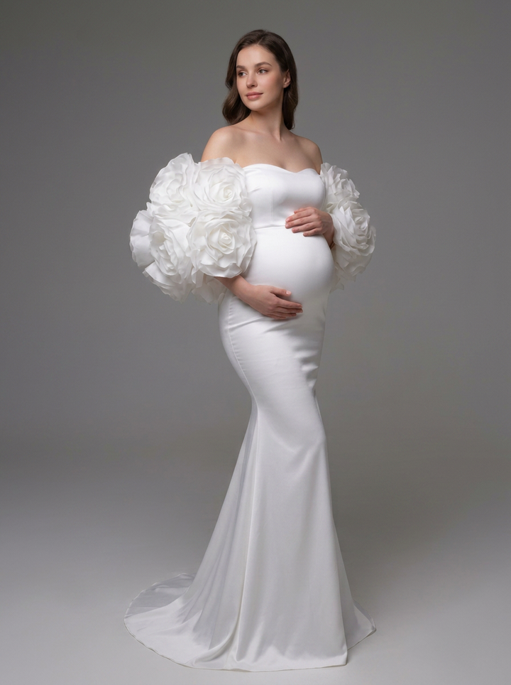 A pregnant model wearing a white off-the-shoulder maternity gown with dramatic, oversized ruffled balloon sleeves and a fitted mermaid silhouette, standing against a neutral gray studio background.