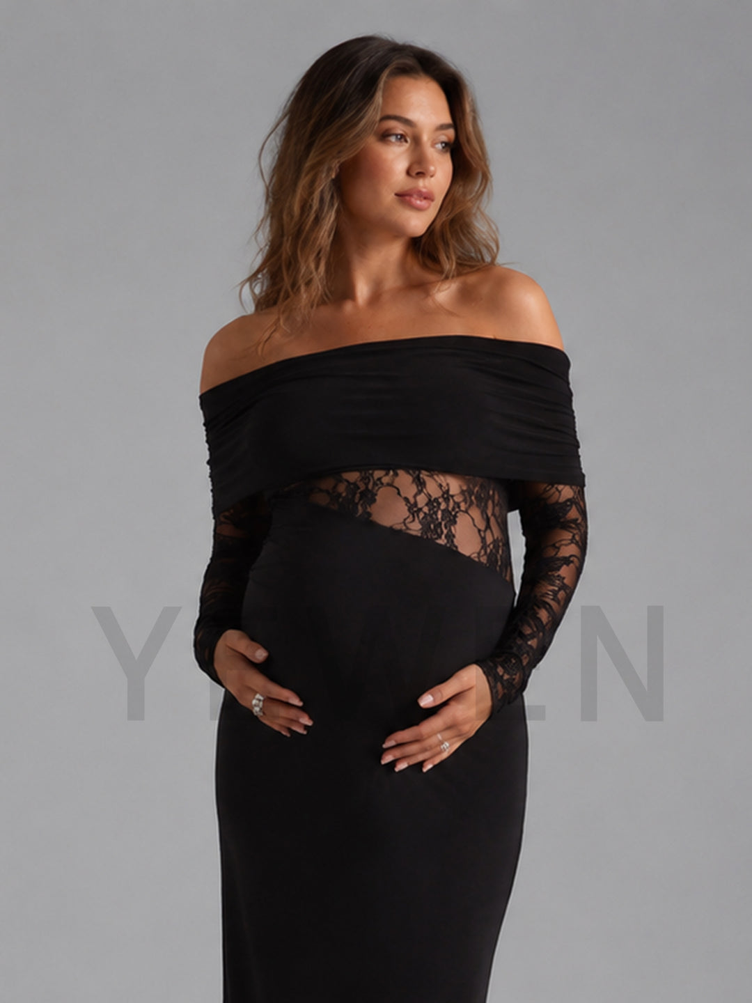 A pregnant model wearing a black off-the-shoulder maternity gown with long sheer lace sleeves and a lace waist panel, posed against a solid gray studio background.