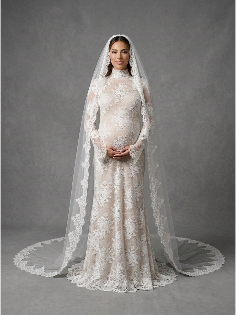 A pregnant model wearing a long-sleeved ivory lace wedding gown with a matching high-neck veil and matching lace earrings, standing against a textured gray studio backdrop.