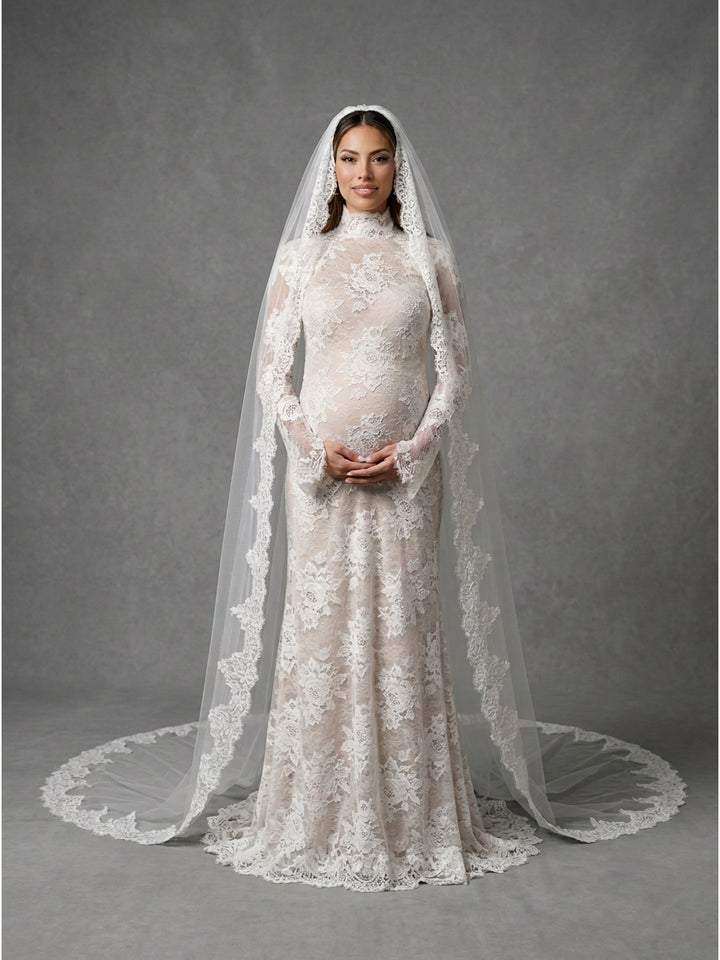 A pregnant model wearing a long-sleeved ivory lace wedding gown with a matching high-neck veil and matching lace earrings, standing against a textured gray studio backdrop.