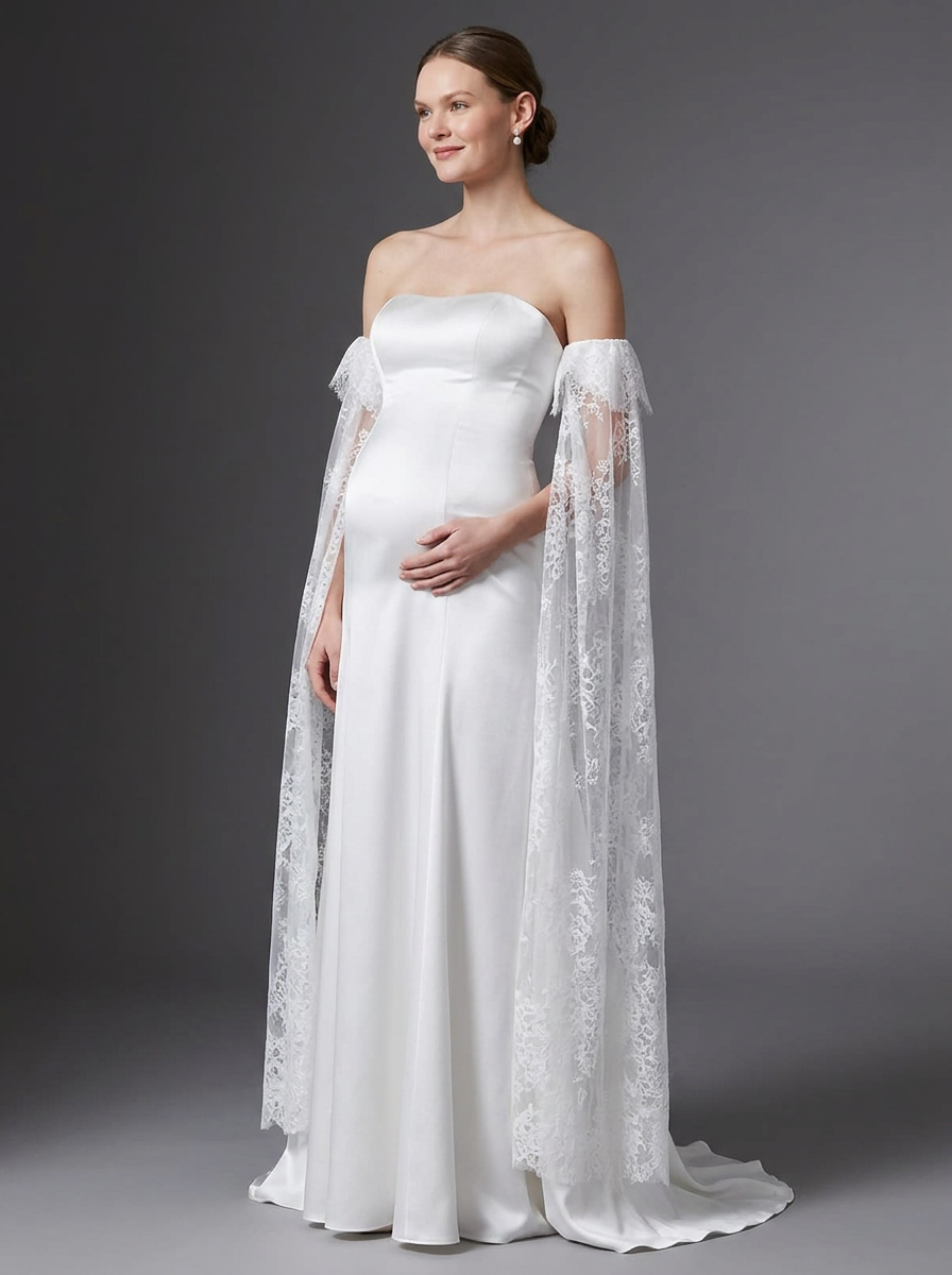A woman modeling a strapless white wedding gown with a long sheer lace cape overlay, photographed against a dark gray studio background.