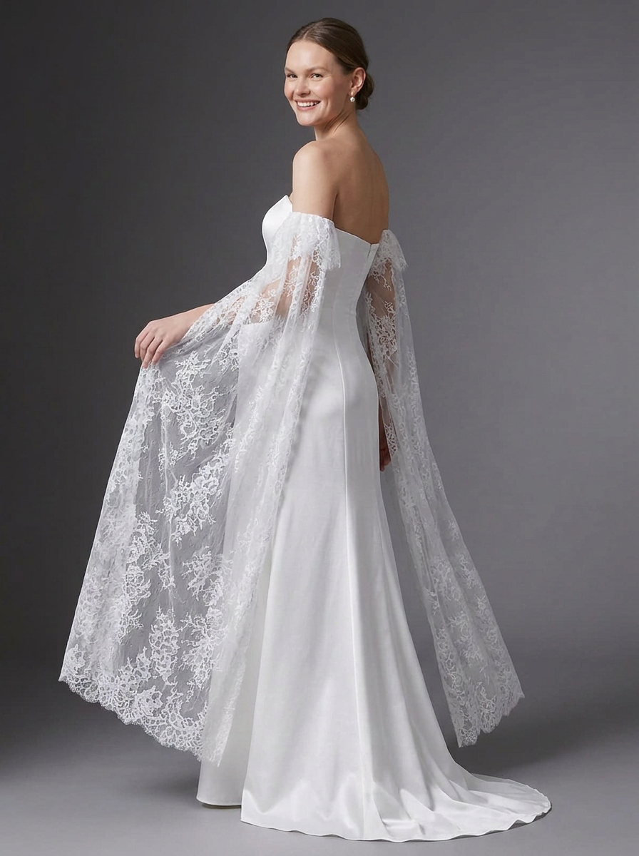 A woman modeling a white off-the-shoulder wedding gown with a sheer lace cape overlay, photographed against a dark gray studio background.