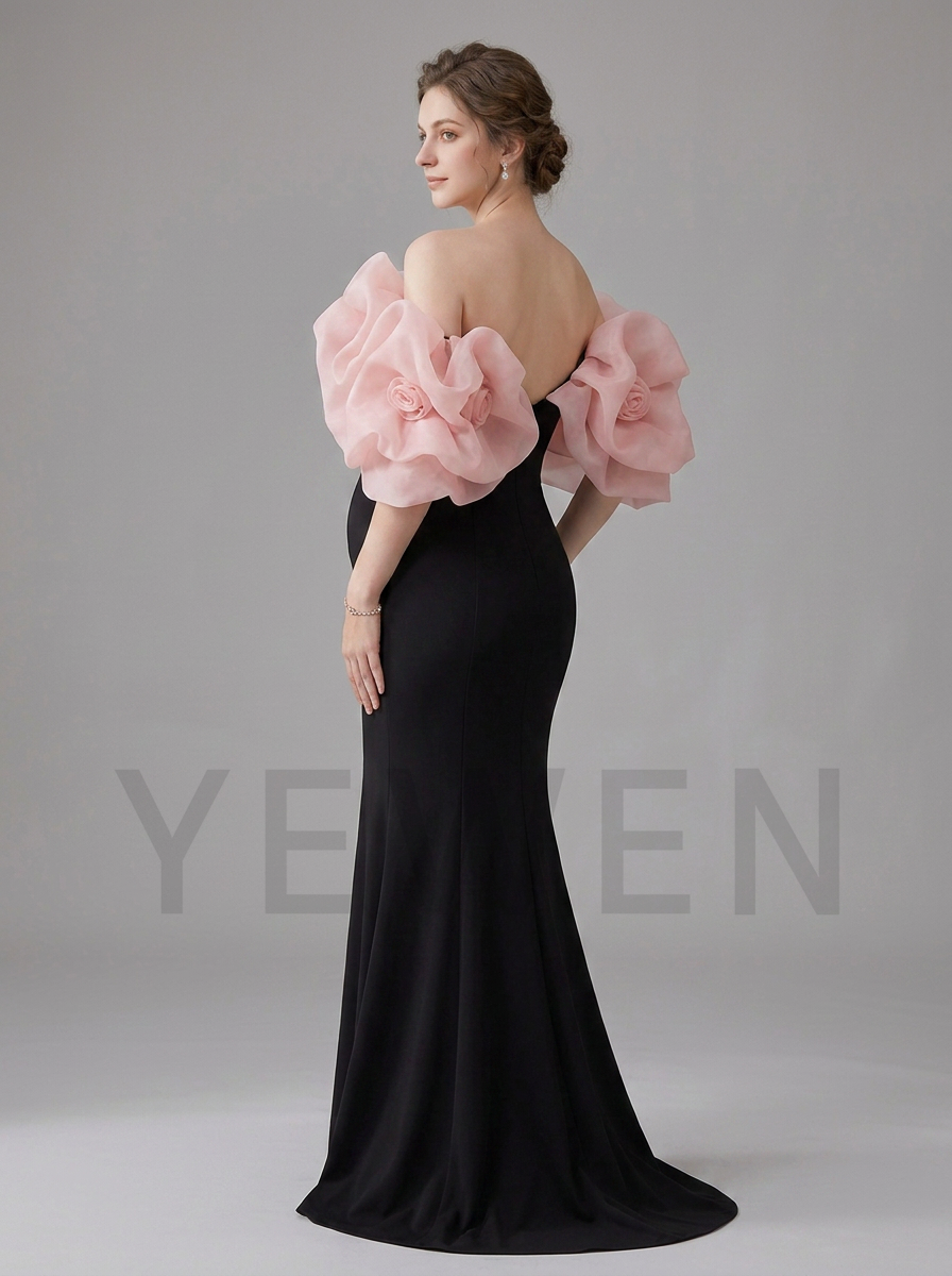 A woman modeling a floor-length black evening gown from the back, featuring dramatic oversized light pink ruffled shoulder details and a low open back, photographed against a plain light gray studio background.