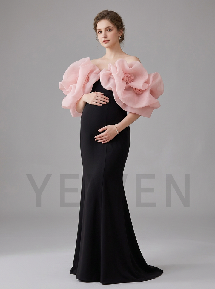 A pregnant model wearing a floor-length black gown with dramatic oversized light pink fabric flower puff sleeves, posed against a neutral gray studio background.
