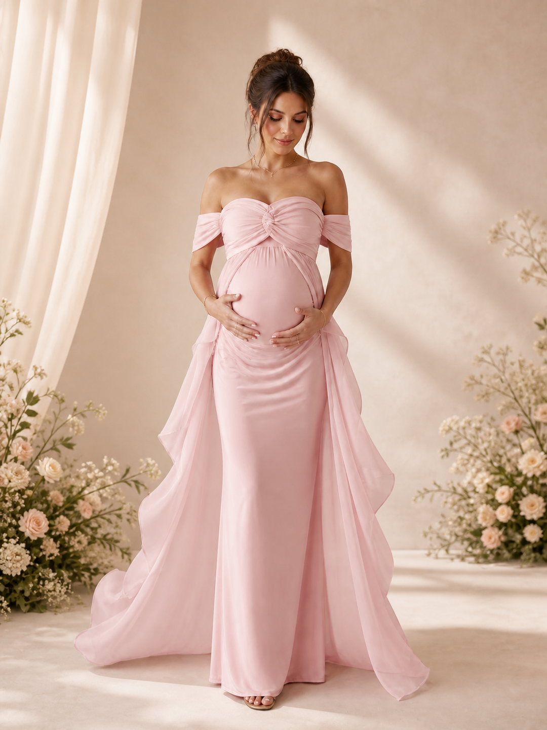 A pregnant woman wearing a light pink off-the-shoulder maternity gown with a ruched sweetheart bodice and flowing skirt, standing in a softly lit studio with floral arrangements and draped fabric.