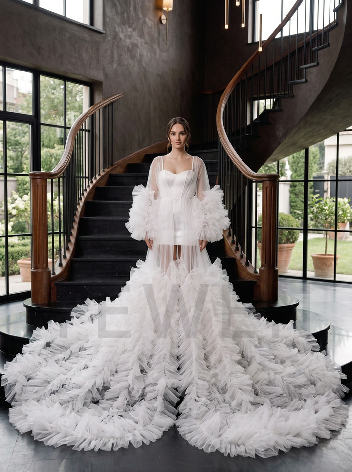 A model stands on a grand staircase wearing a dramatic white wedding gown with sheer long sleeves, voluminous ruffled sleeves, and a long flowing train.