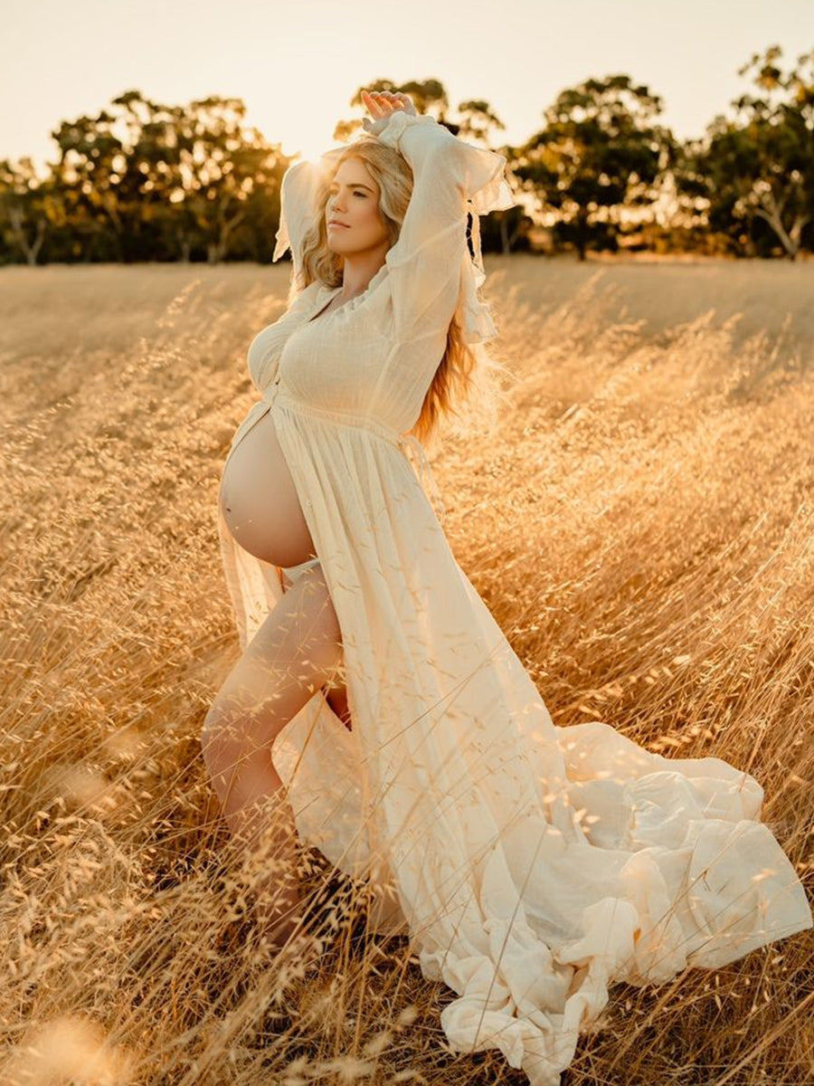 Pregnant woman in a flowing white dress standing in a golden field with trees in the background.