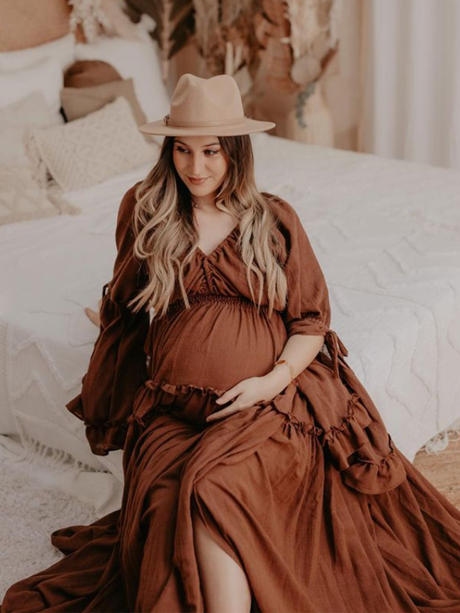 Woman in a brown dress and hat sitting on a bed.