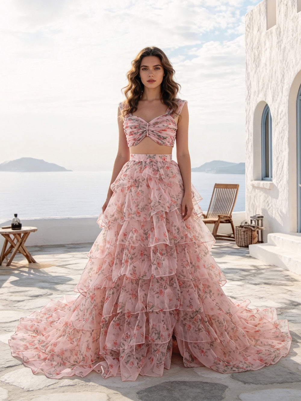 A woman stands on a sunlit stone terrace wearing a pink floral two-piece dress with a fitted crop top and a full layered tulle skirt. The background shows a white building, a chair, and the sea and sky beyond.
