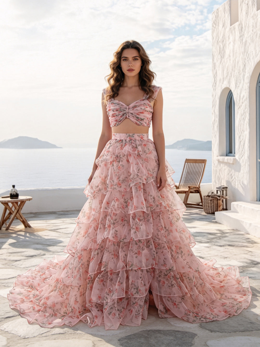 A woman stands on a sunlit stone terrace wearing a pink floral two-piece dress with a fitted crop top and a full layered tulle skirt. The background shows a white building, a chair, and the sea and sky beyond.