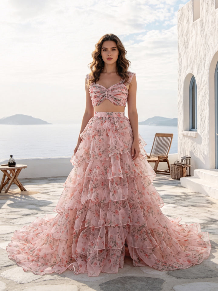 A woman stands on a sunlit stone terrace wearing a pink floral two-piece dress with a fitted crop top and a full layered tulle skirt. The background shows a white building, a chair, and the sea and sky beyond.