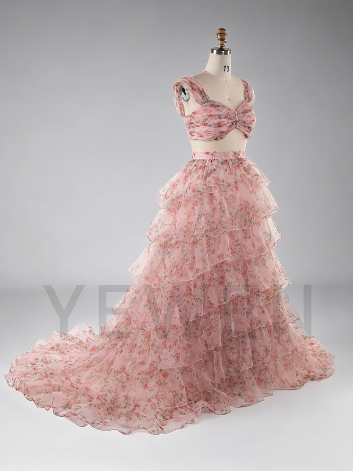 A pink floral two-piece formal dress displayed on a mannequin against a light gray studio background. The set includes a sweetheart-style top with a twisted front detail and a long, layered ruffled skirt.
