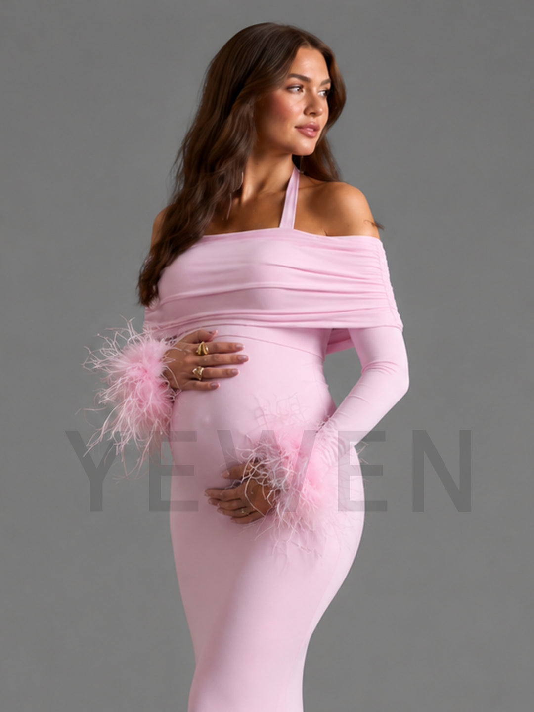 A pregnant woman wearing a light pink off-the-shoulder maternity dress with long sleeves and feathered cuffs, posed against a solid gray studio background.