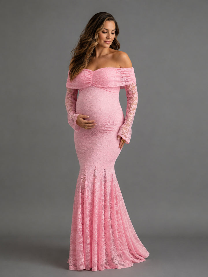 A pregnant woman modeling a light pink off-the-shoulder lace maternity gown with long sleeves and a fitted mermaid silhouette, photographed against a solid gray studio background.