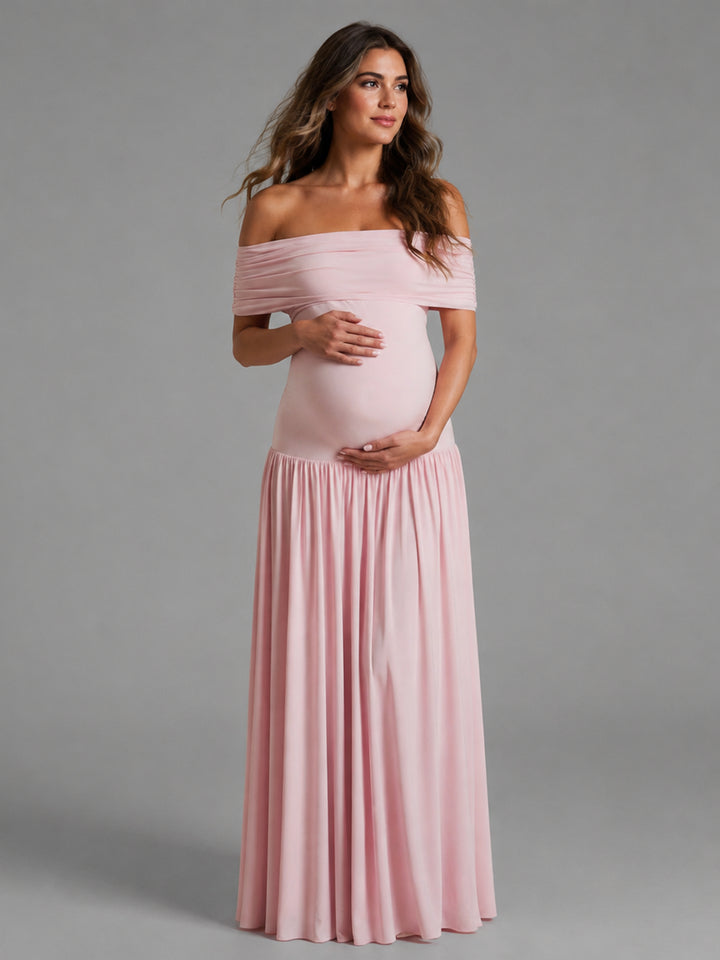 A pregnant woman modeling a light pink off-the-shoulder maternity maxi dress with a ruched bodice and flowing skirt, standing against a solid gray studio background.