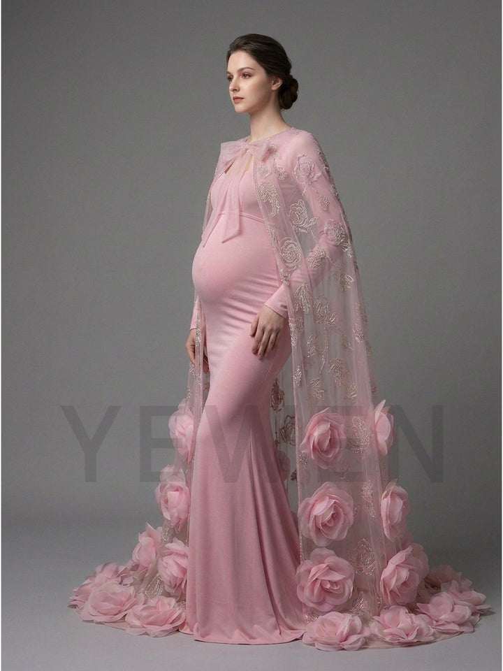 A pregnant model wearing a light pink maternity gown with a sheer floral lace cape and large pink fabric flowers along the hem, photographed against a neutral gray studio background.