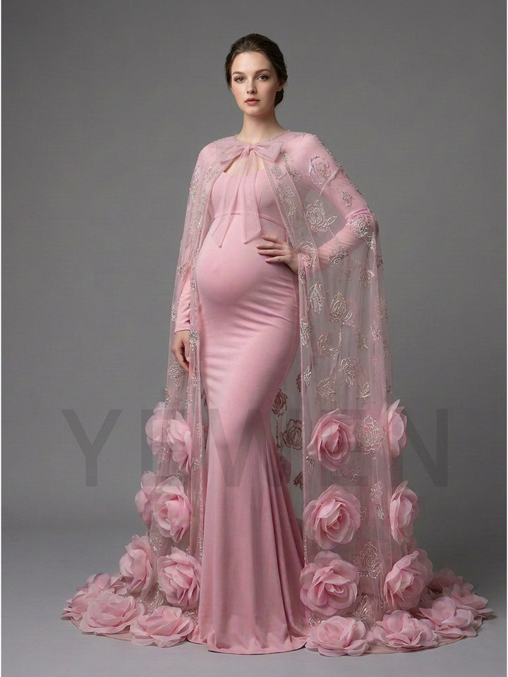 A pregnant model wearing a light pink maternity gown with a matching sheer floral cape, standing against a solid gray studio background.