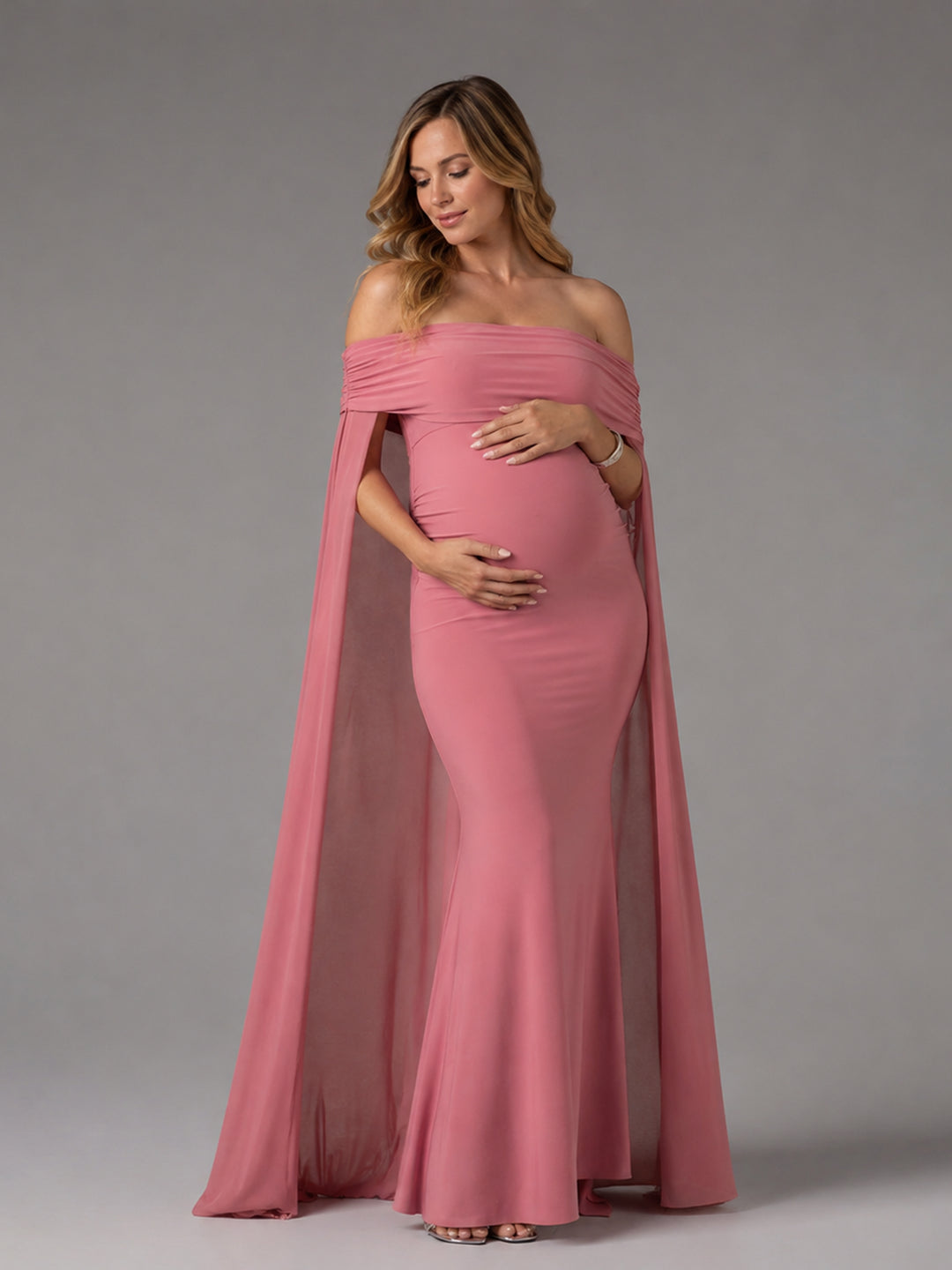 A pregnant woman wearing a dusty rose off-the-shoulder maternity gown with a matching draped cape, standing against a neutral gray studio background.