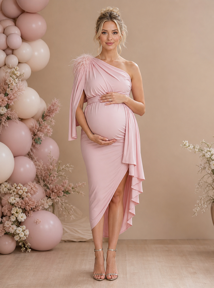 A pregnant woman models a light pink one-shoulder maternity dress with a feathered shoulder detail and a draped ruffled hemline, standing in front of a beige backdrop decorated with pink and white balloons and dried flowers.
