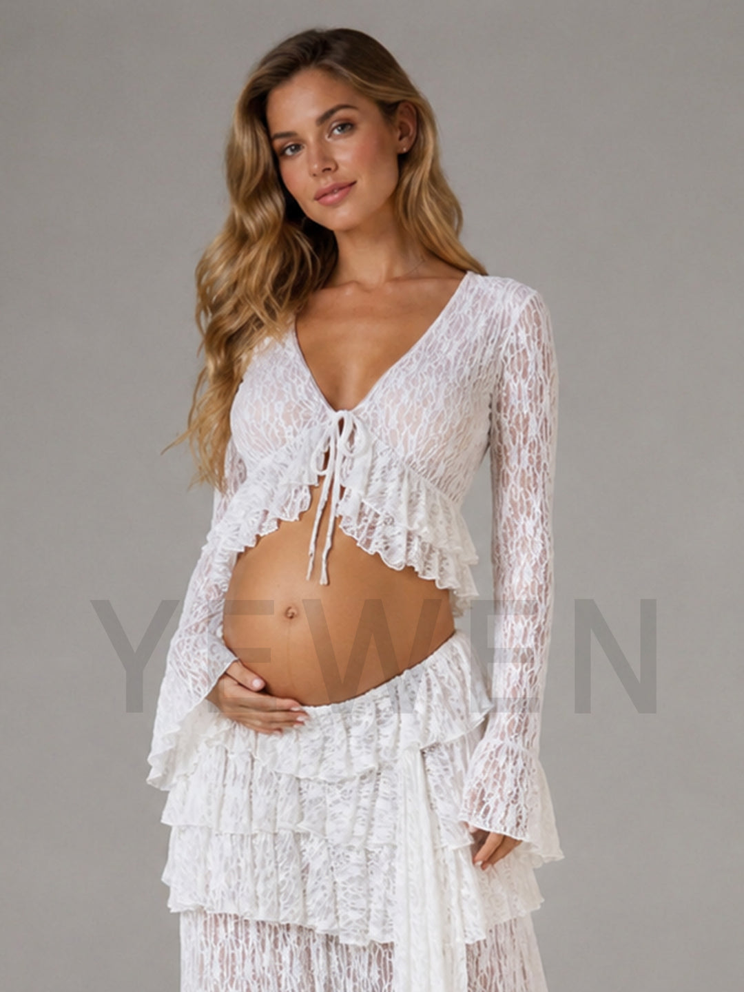 A pregnant woman wearing a white lace long-sleeve crop top with a tie-front detail and a matching ruffled tiered skirt, posed against a solid gray studio background.