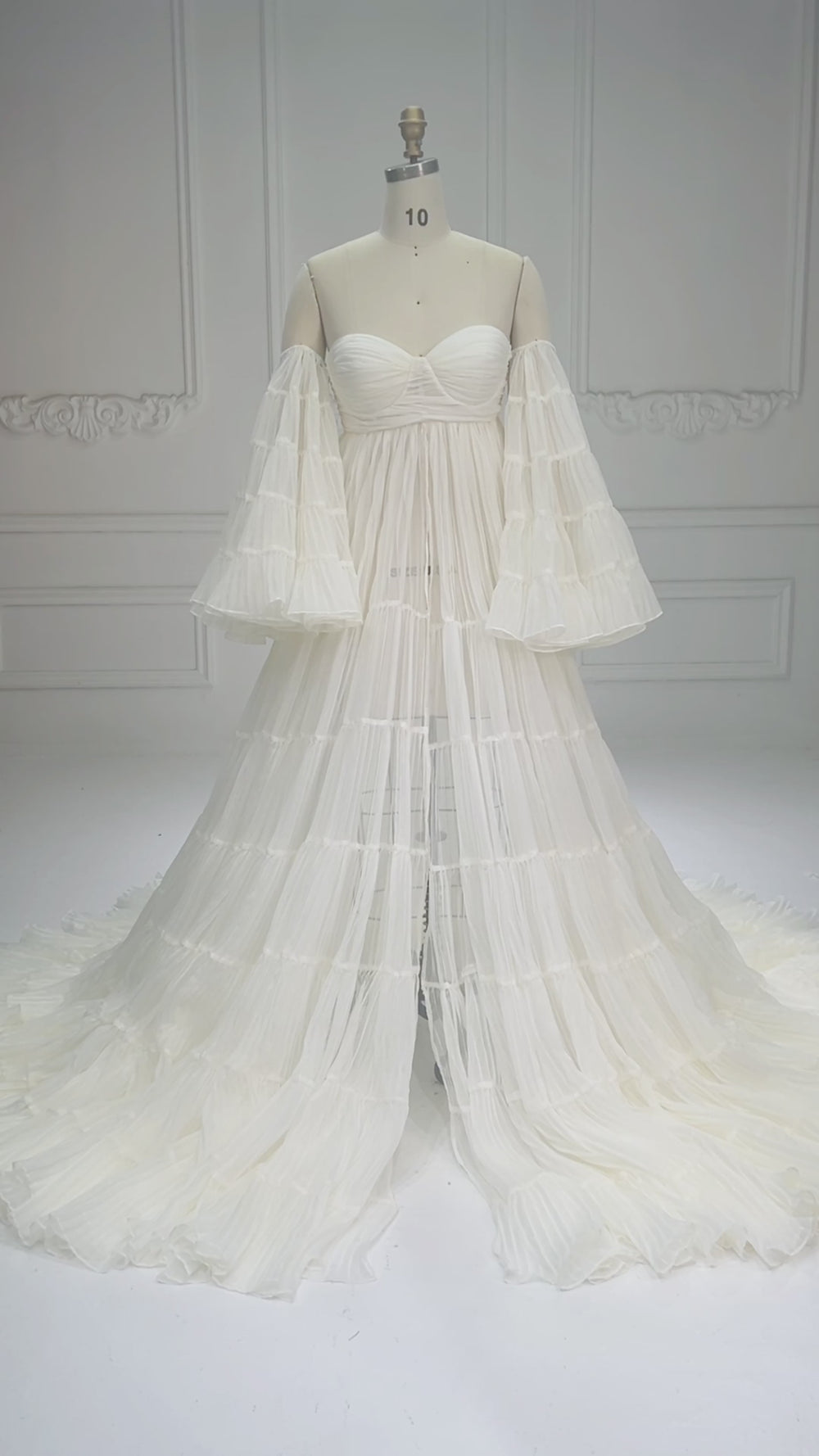 Video showcasing a white pleated organza maternity photoshoot gown with long sheer sleeves a sweetheart neckline and a flowing tiered skirt displayed on a mannequin.