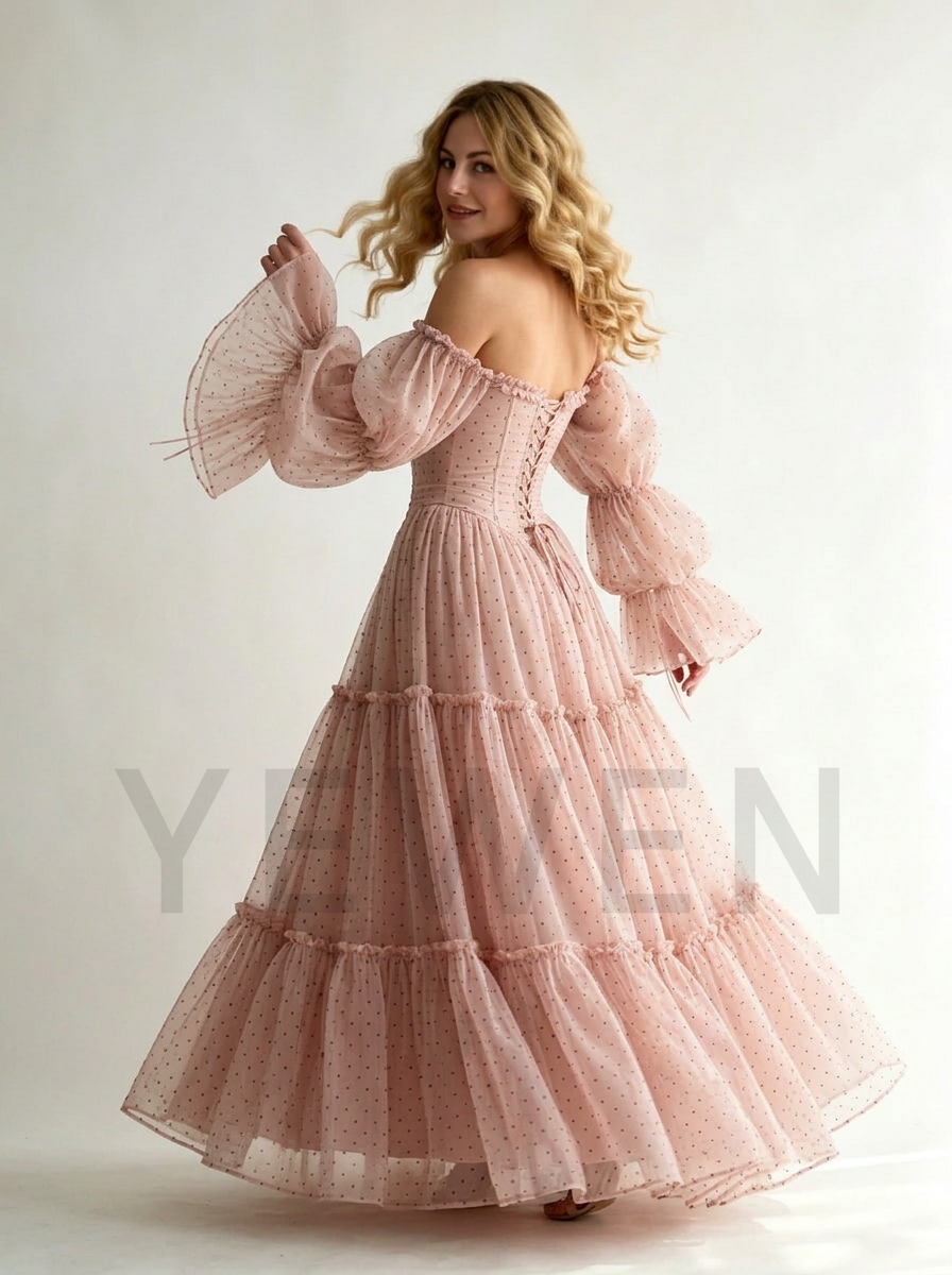 A woman modeling a long, off-the-shoulder pink tulle dress with tiered ruffles and dramatic bell sleeves against a plain light background.