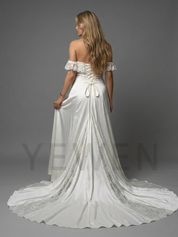 A woman modeling a white off-the-shoulder wedding gown from the back, showing a lace-trimmed back, corset-style waist tie, and a long train on a gray studio background.
