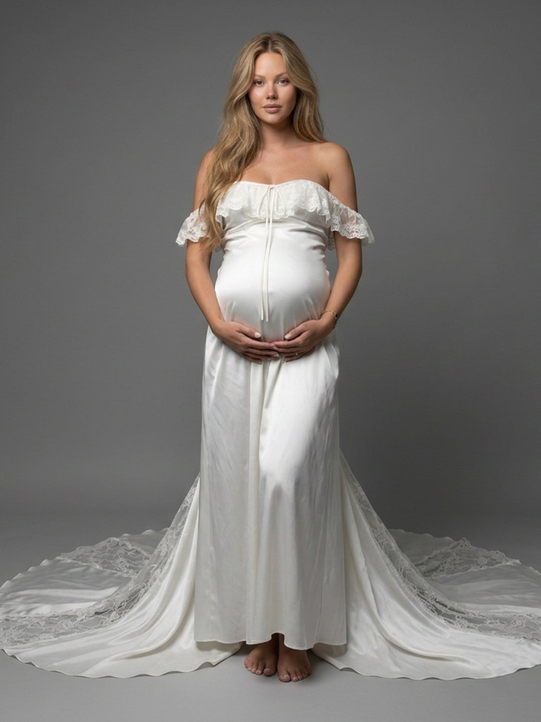 A pregnant woman stands barefoot against a gray studio background wearing an off-the-shoulder white maternity gown with a long train and lace trim.