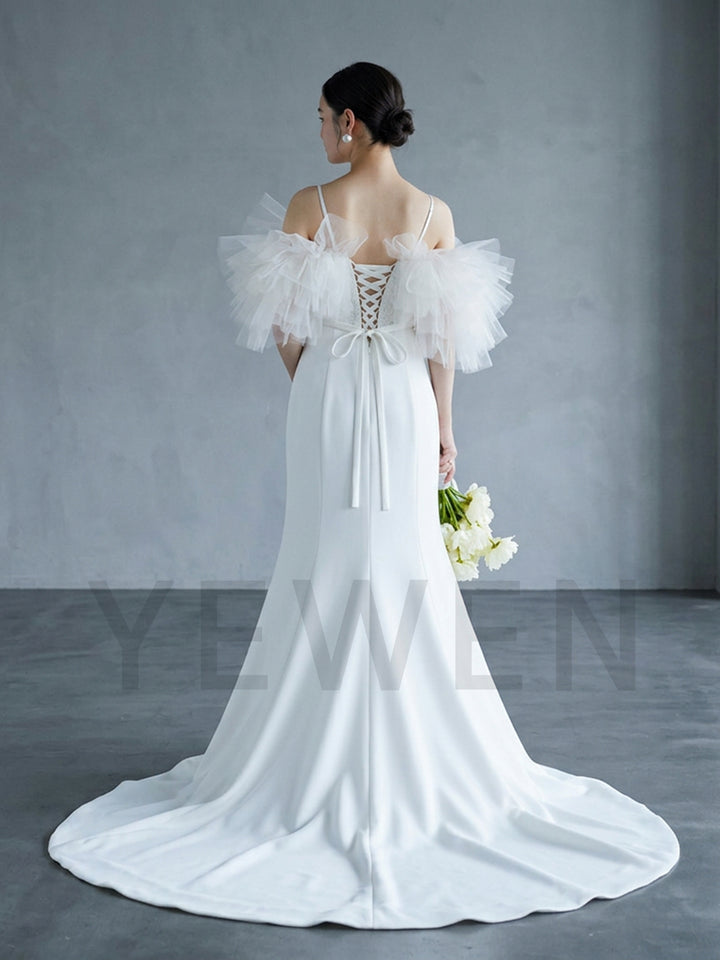 A woman modeling a white off-the-shoulder maternity wedding dress from the back, featuring ruffled tulle sleeves, a lace-up back, and a long train, standing against a gray studio backdrop.