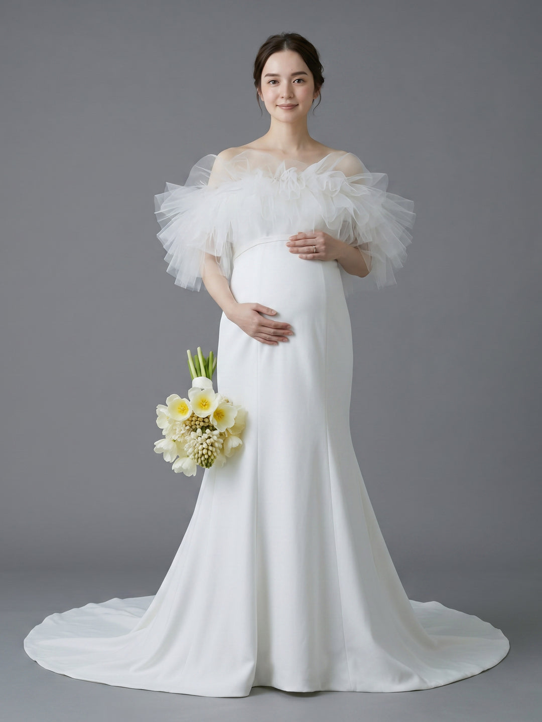 A pregnant woman wearing a white off-the-shoulder maternity wedding dress with dramatic ruffled tulle sleeves and a long train, holding a bouquet of white and yellow flowers against a solid gray studio background.