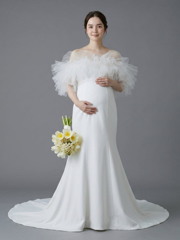 A pregnant woman wearing a white off-the-shoulder maternity wedding dress with dramatic ruffled tulle sleeves and a long train, holding a bouquet of white and yellow flowers against a solid gray studio background.