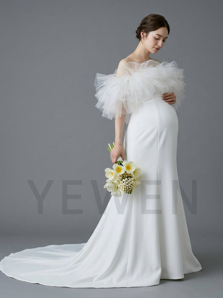 A pregnant woman wearing a white off-the-shoulder maternity wedding dress with a dramatic ruffled tulle sleeve and a long train, holding a bouquet of white flowers against a solid gray studio background.
