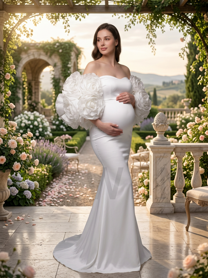 A pregnant woman stands in a sunlit garden wearing a fitted white gown with oversized sculptural puff sleeves and a long train. The setting includes stone arches, flowers, greenery, and a warm outdoor background.