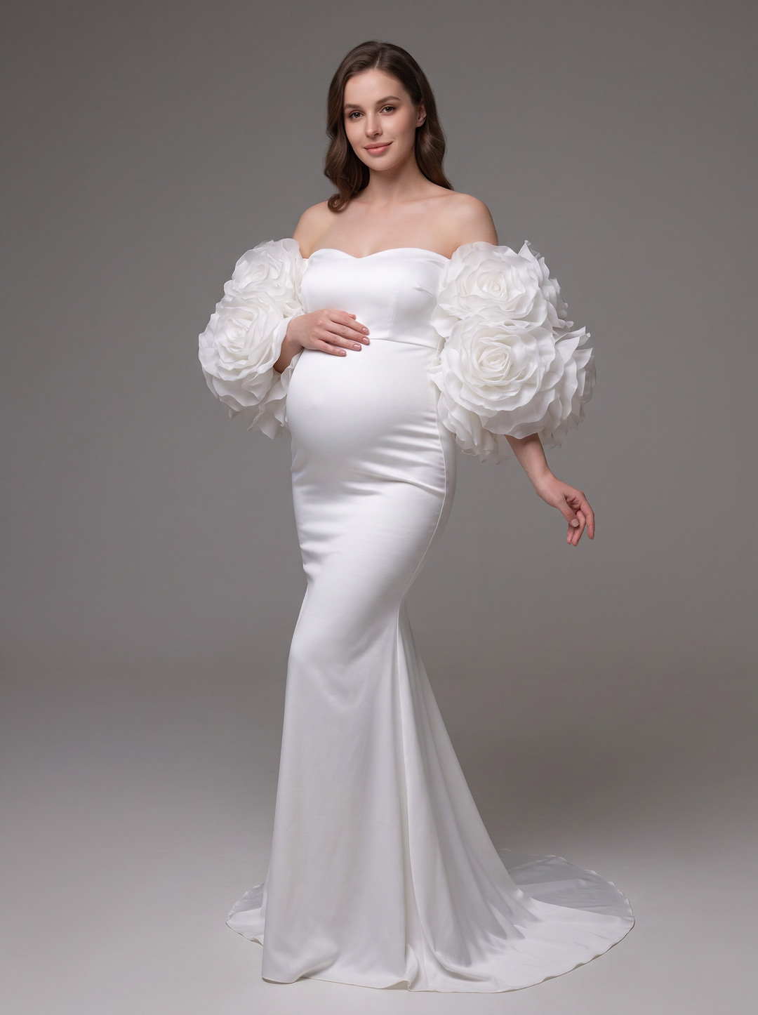 A pregnant model wearing a white off-the-shoulder mermaid gown with oversized 3D fabric roses on the sleeves, standing against a neutral gray studio background.