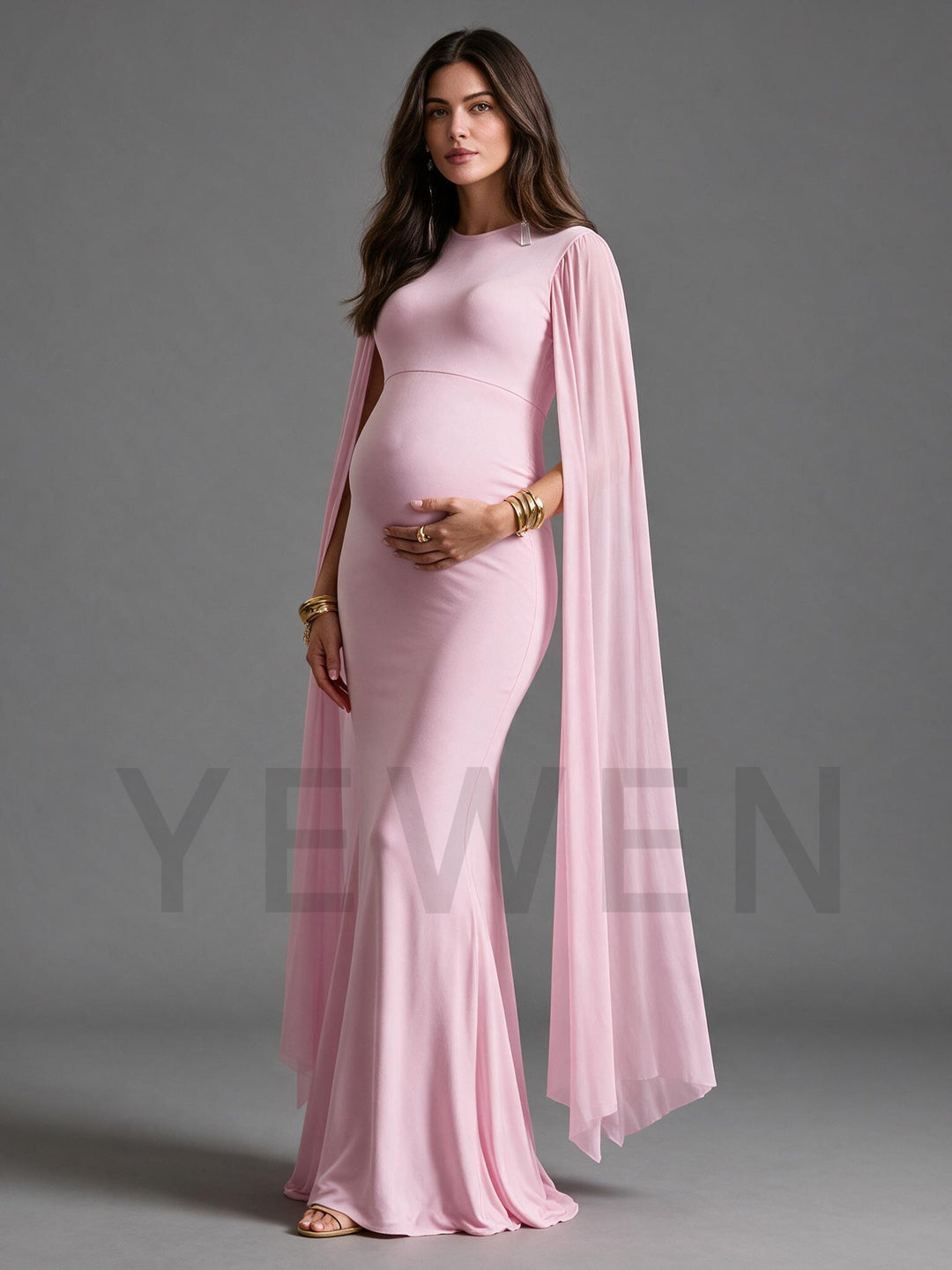 A pregnant woman modeling a light pink maternity gown with a high neckline, fitted silhouette, and a flowing sheer cape-style overlay, posed against a solid gray studio background.