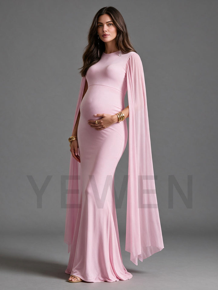 A pregnant woman modeling a light pink maternity gown with a high neckline, fitted silhouette, and a flowing sheer cape-style overlay, posed against a solid gray studio background.
