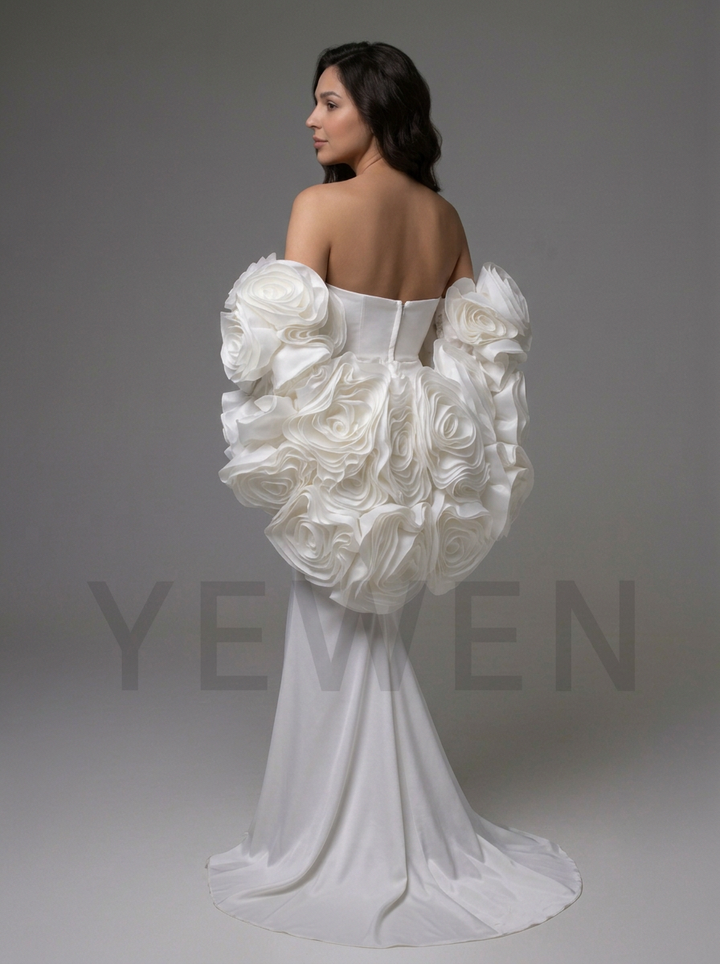 A woman modeling a white off-the-shoulder wedding gown from the back, featuring dramatic sculptural ruffle sleeves and a long train, against a plain gray studio background.