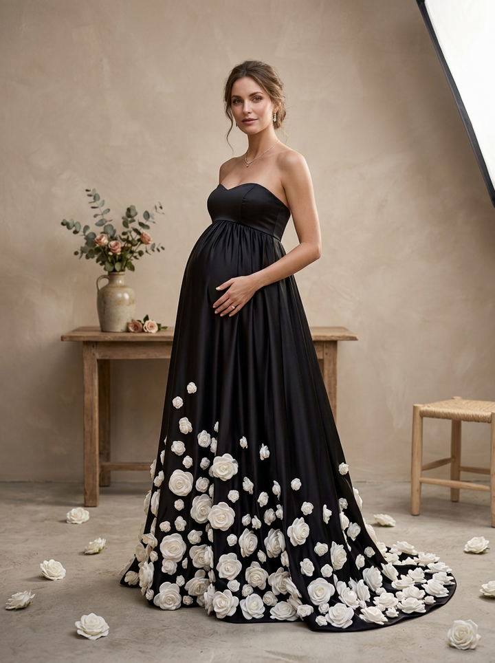 A pregnant woman models a strapless black maternity gown with a sweetheart neckline and a dramatic train covered in large white rose appliqués, set against a warm beige studio backdrop with a wooden table and vase.