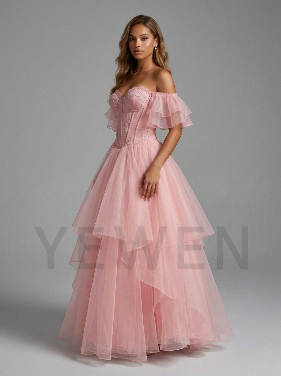 A woman modeling a formal off-the-shoulder pink gown with a structured corset bodice and a layered tulle skirt, photographed against a plain gray studio background.