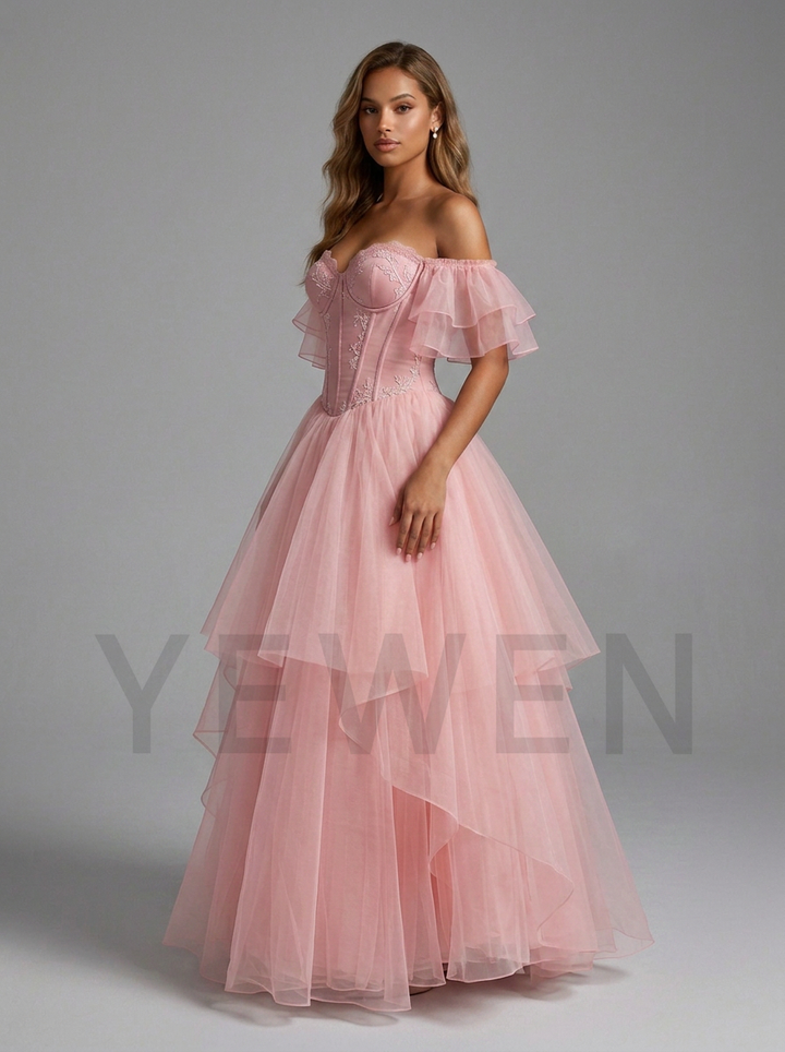A woman modeling a formal off-the-shoulder pink gown with a structured corset bodice and a layered tulle skirt, photographed against a plain gray studio background.