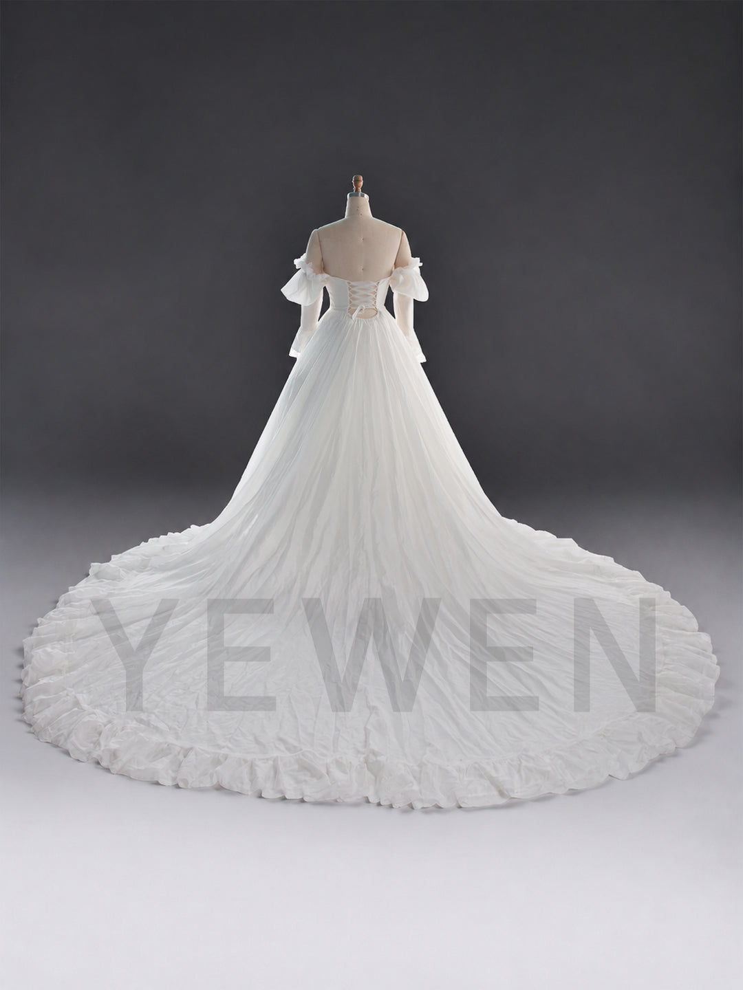 An off-the-shoulder white wedding gown displayed on a dress form mannequin against a dark gray studio background. The gown features a structured bodice, sheer flutter sleeves, and a very large, dramatic ruffled train.