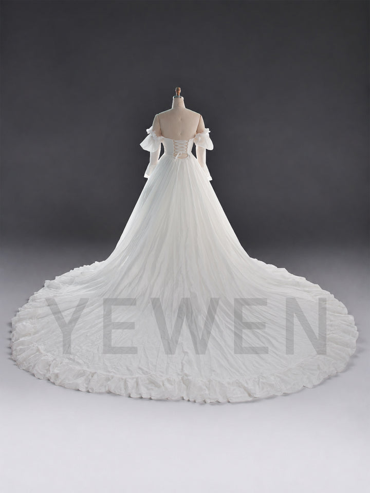 An off-the-shoulder white wedding gown displayed on a dress form mannequin against a dark gray studio background. The gown features a structured bodice, sheer flutter sleeves, and a very large, dramatic ruffled train.