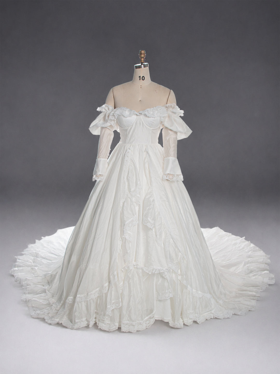 An off-the-shoulder white ball gown with long sheer sleeves and a full tiered skirt, displayed on a dress form against a dark gray studio background.