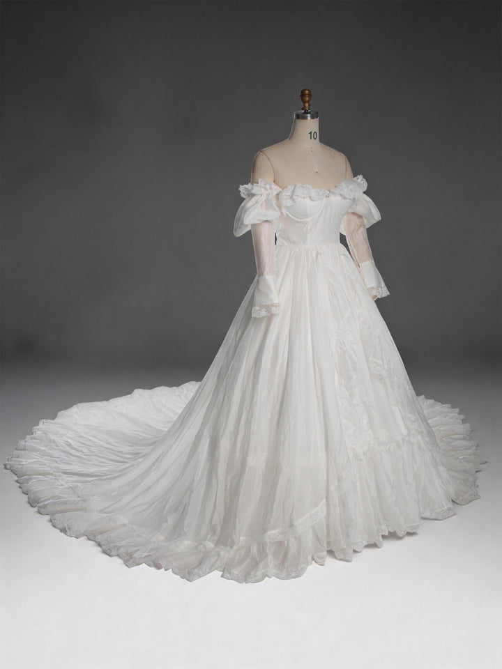 An off-the-shoulder white ball gown with a full skirt and long sheer sleeves, displayed on a dress form mannequin against a dark gray studio background.