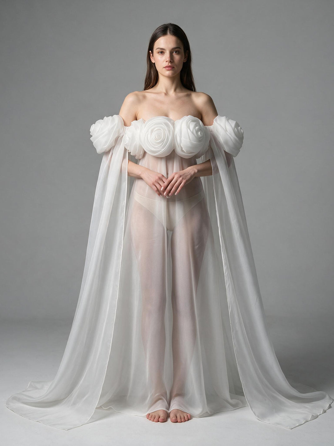 A model stands barefoot against a neutral gray studio background wearing a white off-the-shoulder gown with dramatic sculptural floral shoulder details and a long flowing sheer cape.