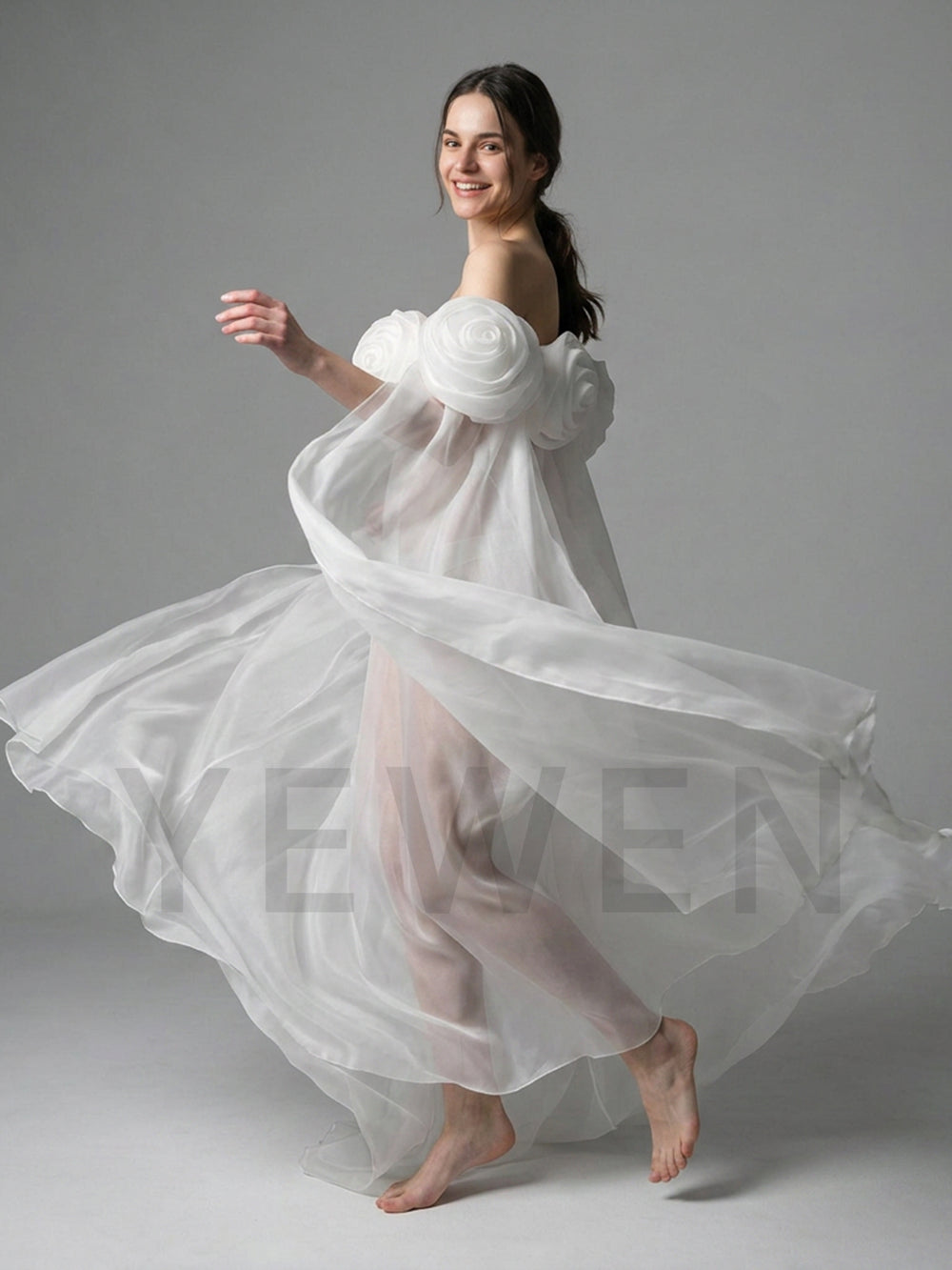 A woman in a flowing white sheer dress with oversized floral shoulder details and bare feet, captured mid-motion against a plain light gray studio background.