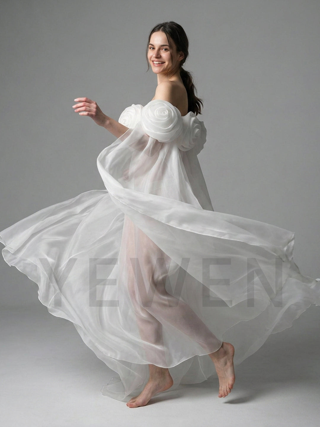 A woman in a flowing white sheer dress with oversized floral shoulder details and bare feet, captured mid-motion against a plain light gray studio background.
