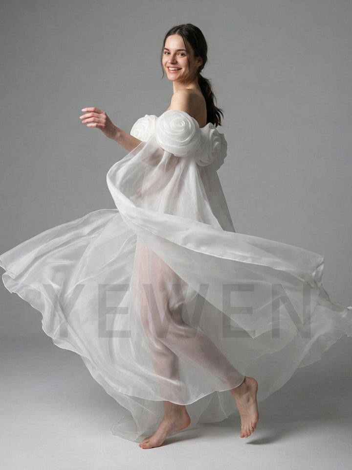 A woman in a flowing white sheer dress with oversized floral shoulder details and bare feet, captured mid-motion against a plain light gray studio background.