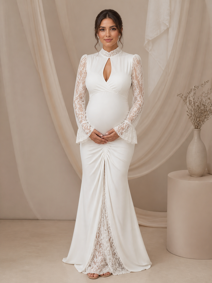 A pregnant model wearing a white maternity gown with long lace sleeves, a high mock neck, and a gathered front, standing in a studio with draped beige fabric and a side table with a vase.