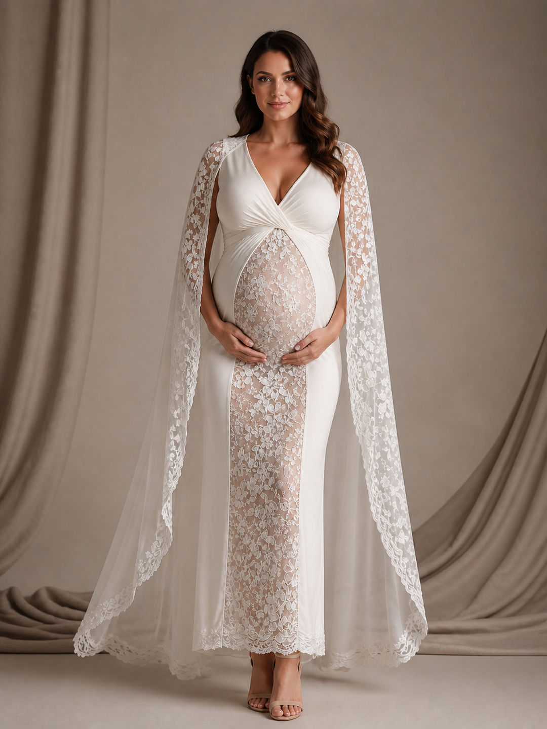 A pregnant woman models a white maternity gown with a lace overlay and a matching sheer lace cape, set against a neutral draped studio backdrop.